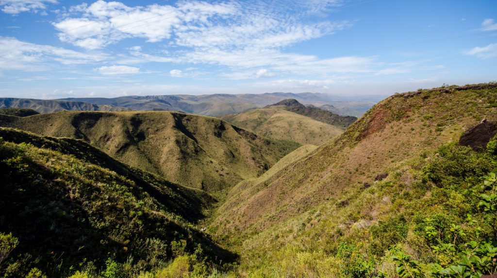 A imagem mostra uma vista panorâmica do Parque Estadual da Serra da Rola Moça, em Minas Gerais, com grandes montanhas e um céu aberto.